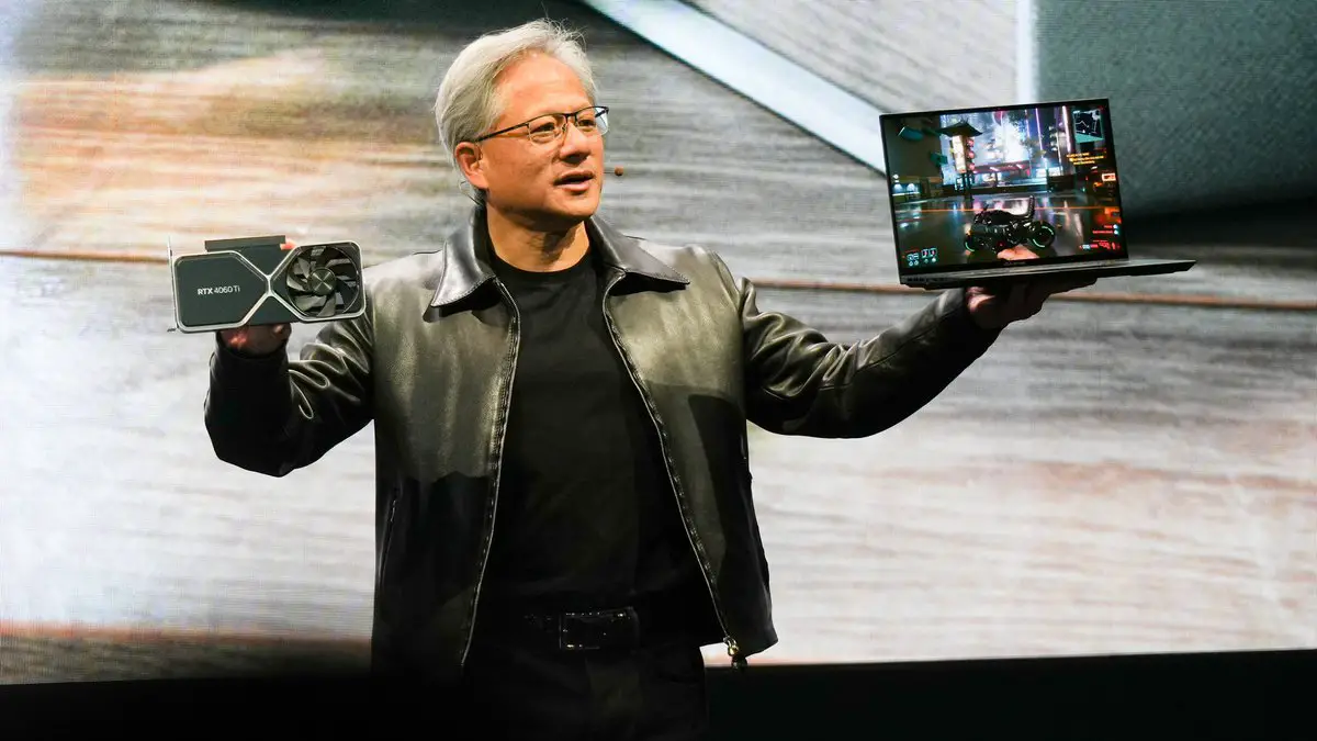 A man holding a graphics card in one hand and a laptop displaying a video game on its screen in the other hand, standing against a blurred background. He is wearing a black shirt and a leather jacket, with glasses and short gray hair.