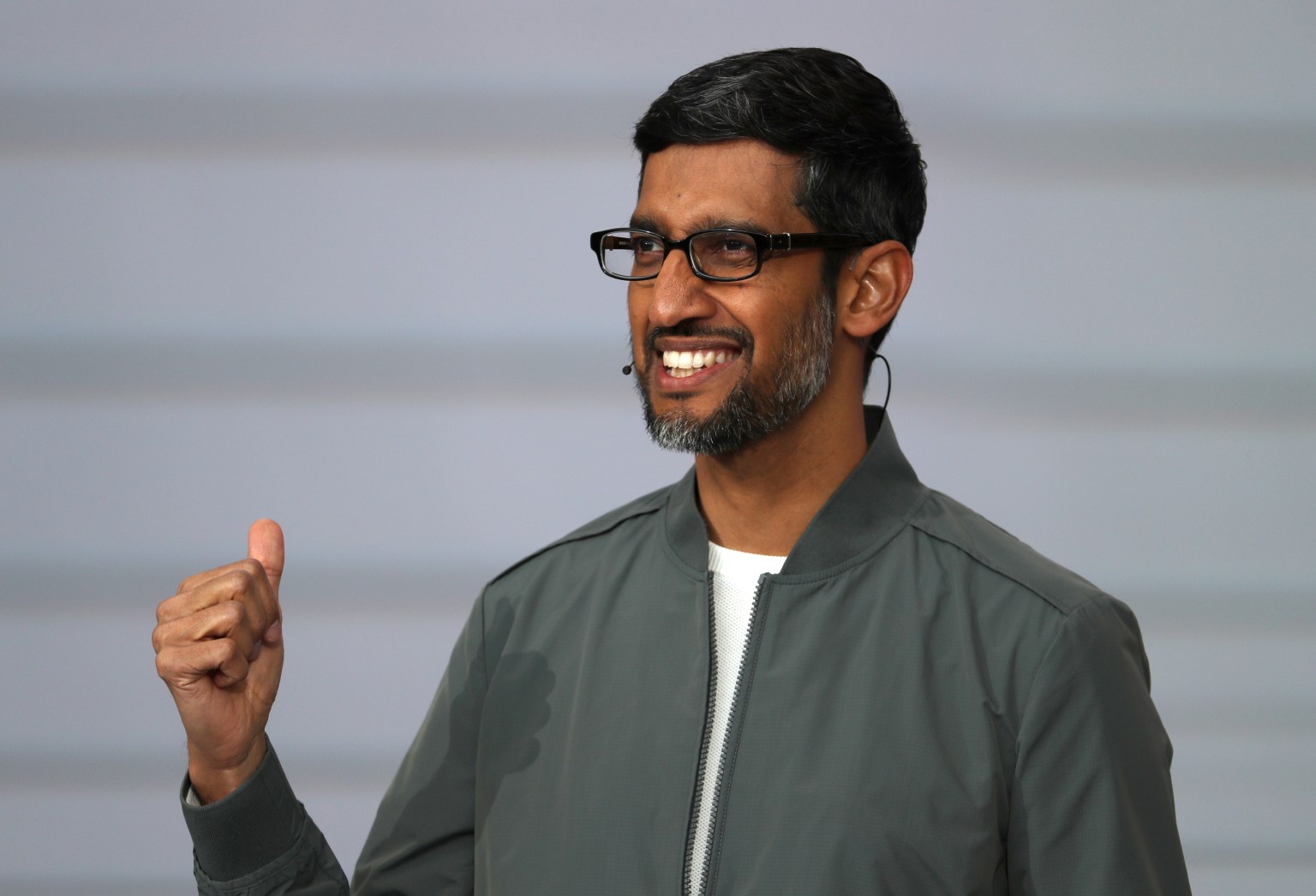 A smiling man wearing glasses and a gray jacket stands with his right hand raised in a thumbs-up gesture, against a neutral background.