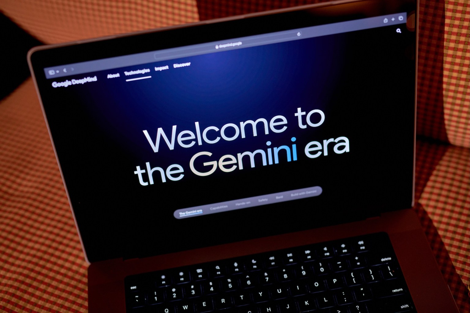 A laptop displaying a dark screen with the text "Welcome to the Gemini era" prominently featured in large, colorful fonts. The background includes a blurred pattern of woven fabric, suggesting a cozy indoor setting.