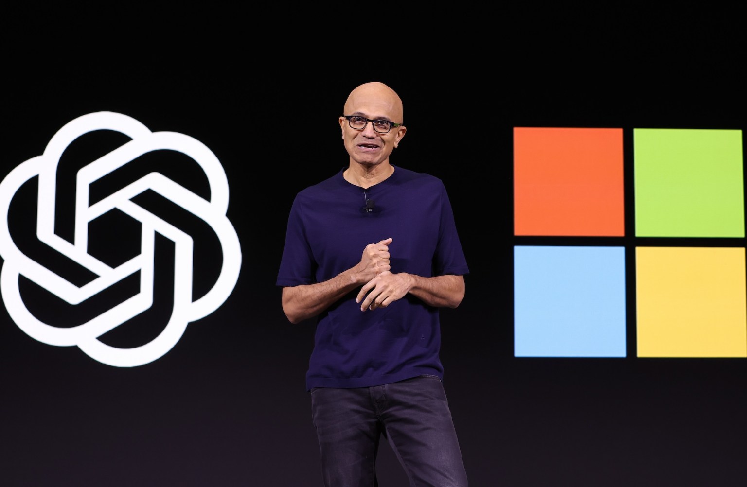 A speaker in a navy t-shirt stands on stage in front of a large screen displaying the OpenAI logo on the left and the Microsoft logo on the right, gesturing with their hands as they present.