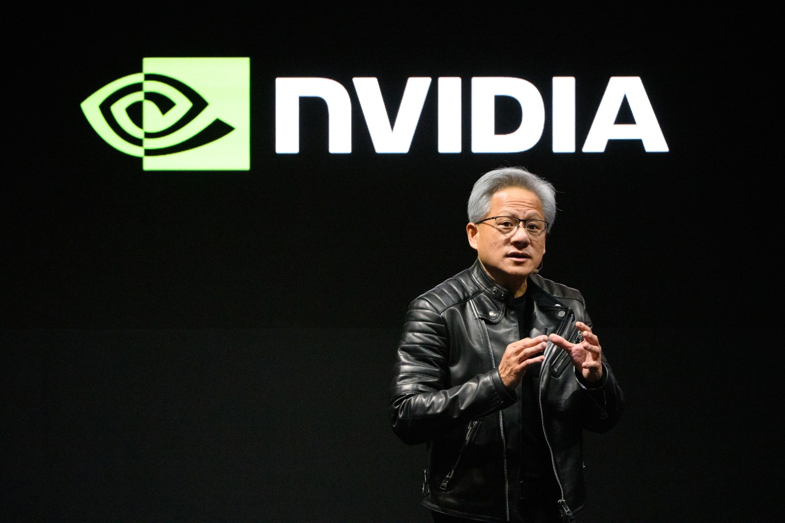 A speaker in a black leather jacket gestures while presenting in front of a large Nvidia logo displayed on a dark background. The speaker appears engaged and enthusiastic, conveying important information to the audience.