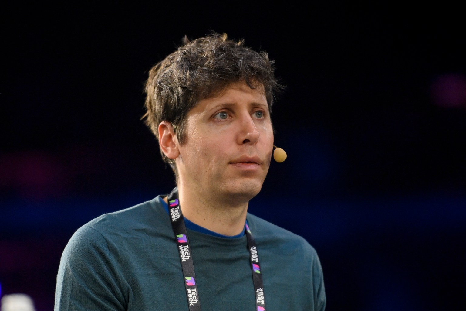 A person with wavy dark hair is wearing a blue shirt and a microphone headset, looking thoughtfully into the distance. They have a tech conference lanyard around their neck, and the background features blurred colorful lights, suggesting an event setting.