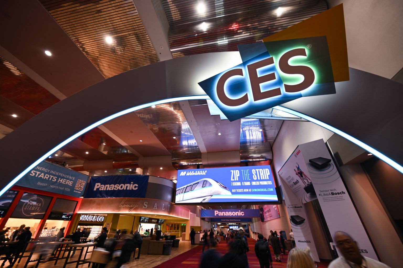 Entrance to CES event featuring a large "CES" sign over an archway. The area is bustling with people, and there are banners for Panasonic and other exhibits. Bright lights and a modern interior set the atmosphere of a high-tech conference.